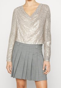 Zkrácený dlouhý rukáv top v metalické béžové barvě se zlacenými flitry, s výstřihem do V. V kombinaci se šedou kostkovanou skládací sukní. Hladká textura, lesklý povrch.