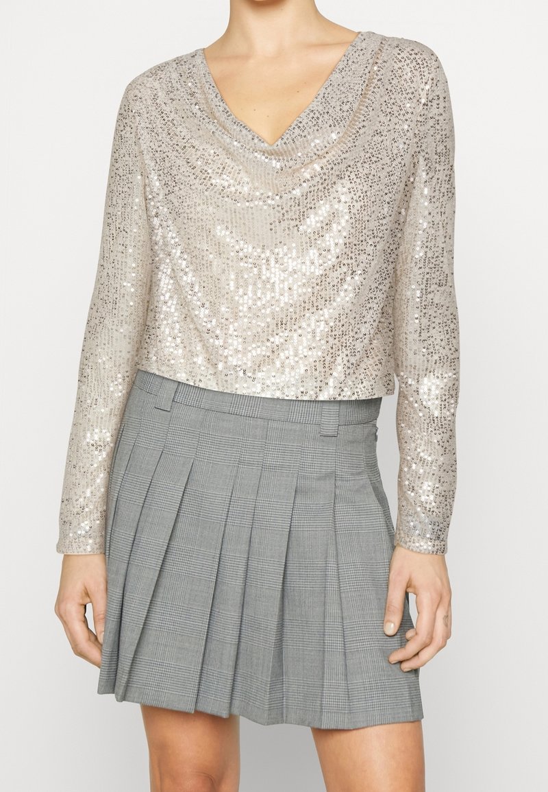 Zkrácený dlouhý rukáv top v metalické béžové barvě se zlacenými flitry, s výstřihem do V. V kombinaci se šedou kostkovanou skládací sukní. Hladká textura, lesklý povrch.