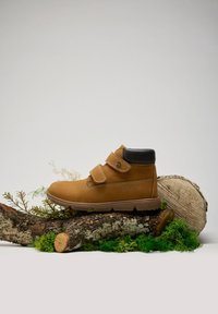 Bottine en daim marron avec des accents en cuir noir, double bandes Velcro, semelle en caoutchouc et texte en relief sur le côté. Positionnée sur une bûche.