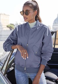 Grey bomber jacket with a zip closure, ribbed cuffs, and collar. Features a side pocket with gold accents. Worn over a light top.