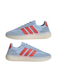 Svijetloplavi tenisici s crvenim trostrukim prugama, detaljima od antilopa, bijelim prozirnim debelim đonovima i vezicama prikazani s lijeve i desne strane.