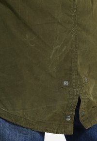 Olivgrön jacka i slitstarkt tyg, med rundad nederkant, tryckknappar och subtil textur med svaga märken.