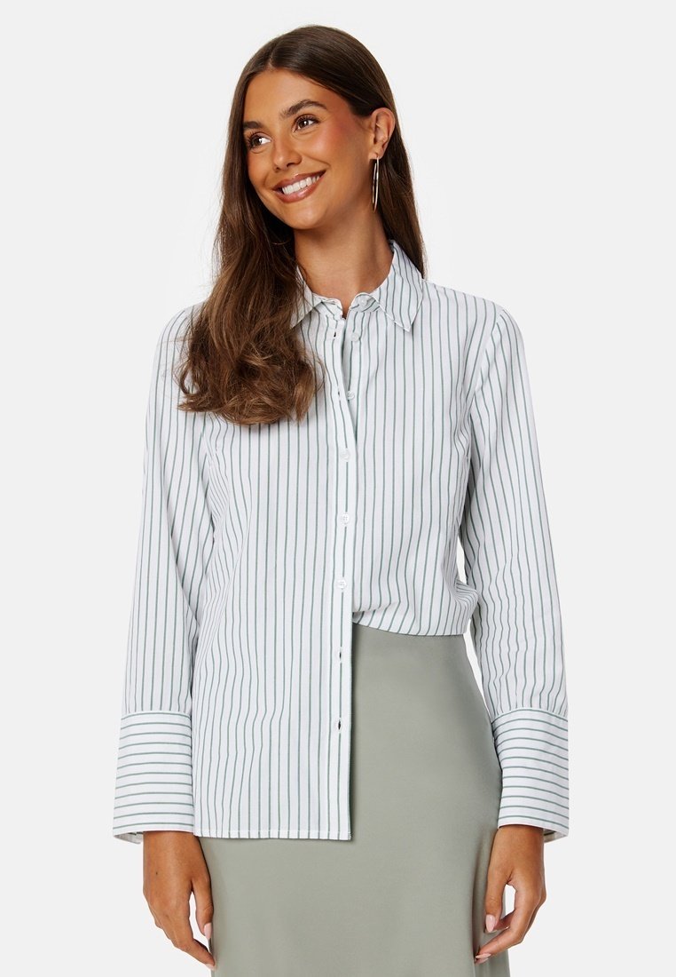 Camicia bianca con bottoni a righe verdi verticali, maniche lunghe e orlo curvo. Abbinata a una gonna verde chiaro. Tessuto morbido.
