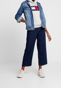 Chaqueta de denim en azul claro, sudadera gris con logo, jeans oscuros de pierna ancha, y zapatillas blancas con acentos rojos. Ajuste relajado, estilo casual.