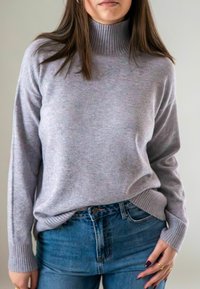 Vrouw in een lichtpaarse coltrui die is gestopt in hoge taille blauwe jeans, met ringen om haar rechterhand.