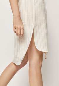 Biała sukienka z pionowymi złotymi paskami, długości midi z bocznym rozcięciem, wykonana z lekkiego materiału, z prostym wykończeniem i minimalnymi detalami.