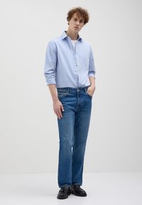 Helles blaues Hemd, weißes Unterhemd, blaue Jeans und schwarze Slipper. Slim-Fit mit hochgekrempelten Ärmeln und normalen Jeans-Taschen.