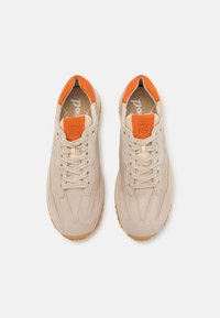 Beige sneakers med oransje detaljer, med en teksturert semsket skinn- og skinn-overdel, snøring, og en gummisåle med mønstrede dekk.