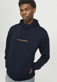 Felpa blu navy in morbido tessuto, con tasca a marsupio, cappuccio con cordino e logo arancione "JACK & JONES" sul petto.