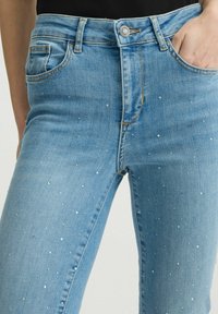 Jeans in denim azzurro chiaro con vestibilità slim, caratterizzati da applicazioni di borchie argentate e un classico design a cinque tasche. Chiusura con bottone in vita.