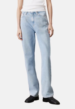 Personne portant un jean droit bleu clair, un haut blanc sans manches et des chaussures noires à bout carré, posant devant un fond uni.