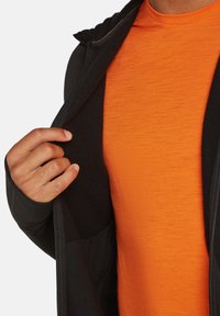Zwarte zip-up hoodie met een zachte binnenvoering, gedeeltelijk geopend om een gestructureerd oranje shirt eronder te onthullen. Hand geplaatst op de hoodie.