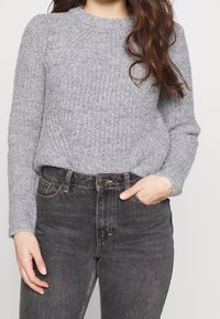 Pull gris en maille avec texture côtelée, col rond et manches longues, associé à un jean en denim foncé taille haute avec poches frontales.