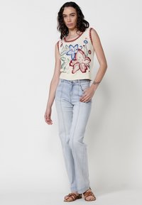 Top sin mangas de punto color blanco roto con bordado floral, combinado con jeans de mezclilla de lavado claro con costuras laterales. Llevado con sandalias marrones con tiras.