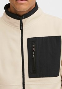 Chaqueta de forro polar beige con un bolsillo en el pecho de nailon negro. Presenta un cuello alto y cierre delantero, exhibiendo una mezcla de texturas.