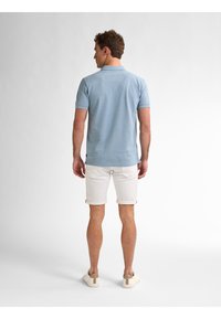 Polo de color azul claro con mangas cortas, combinado con shorts blancos enrollados. El modelo lleva zapatillas beige con un acento en el talón de un color contrastante.