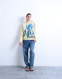 Νεαρός με μαλλιά τραβηγμένα προς τα πίσω, που φοράει μπεζ μακρυμάνικο πουκάμισο με μπλε ανθισμένο σχέδιο, χαλαρά μπλε τζιν και μαύρα σανδάλια, στέκεται μπροστά σε λευκό τοίχο.