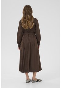 Brown long-sleeved dress with a cinched waist, pleated back, and ankle-length hem. Paired with black and white striped flats.
