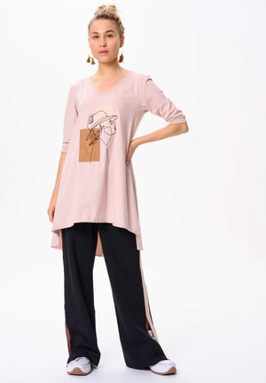 Roze tuniek met een V-hals en driekwart mouwen, voorzien van een lijntekening aan de voorkant. Gepaarde met zwarte wijde broek met zijsplitten.