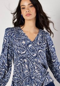 Blau-weiße Bluse mit Paisley-Muster, V-Ausschnitt, langen Ärmeln und gefälteltem Schulterdetail. Der Stoff wirkt leicht und hat eine glatte Textur.