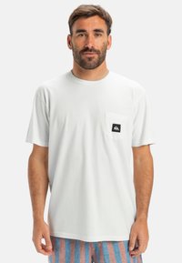 Bílá bavlněná tričko s krátkým rukávem, kulatým výstřihem a přední kapsou s malou černou nášivkou loga. Pozadí je jednobarevné.