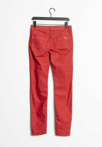 Červené džíny z denimu s úzkým střihem, které mají dvě zadní kapsy, pevný pas a hladkou texturu. Visí na černém plastovém ramínku.