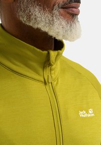 Groene fleece jas met ritssluiting en hoge kraag. Bevat vlakke naden, textuurstof en een wit logo op de linker borst.