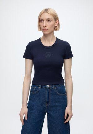 Frau mit kurzen blonden Haaren trägt ein eng anliegendes marineblaues Kurzarm-T-Shirt und hoch sitzende blaue Jeans vor einem schlichten weißen Hintergrund.