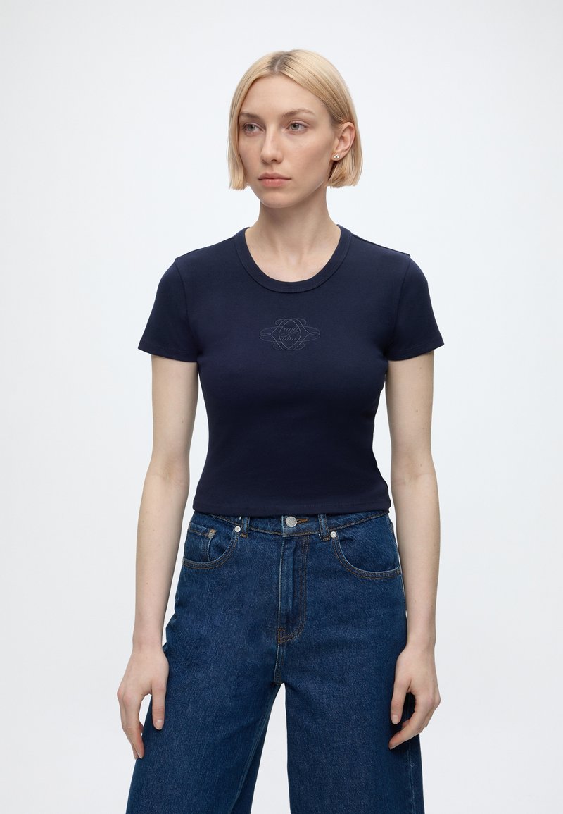 Femme aux cheveux blonds courts portant un t-shirt bleu marine ajusté à manches courtes et un jean bleu taille haute sur un fond blanc uni.