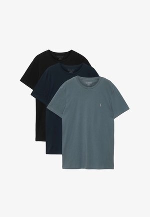 Mann steht, trägt ein schlichtes dunkelblaues T-Shirt, schwarze weit geschnittene Jeans und schwarze Schuhe vor einem einfarbigen hellgrauen Hintergrund.