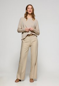 Beige stickad tröja med ribbade manschetter, i kombination med ljusbeiga vida byxor, accentuerad av ett bälte. Slät textur och avslappnad passform.