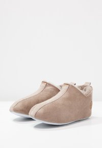 Beige semskede tøfler med mykt, fluffy fôr, buet design og kontrastfarger i sømmen. Har en liten logo på siden.