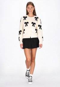 Fiatal nő sétál, fehér kardigánt visel fekete masnikkal, fekete pliszírozott szoknyát, fehér zoknit és fekete sportcipőt egy fehér háttér előtt.