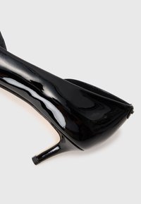 Glänzender schwarzer Lacklederschuh mit hohen Absätzen und einem kurzen, schlanken Absatz auf einem schlichten hellen Hintergrund.