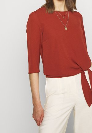 Femme portant un haut couleur rouille à manches trois-quarts avec un nœud sur le côté et un pantalon blanc taille haute, portant deux colliers pendentifs en or.