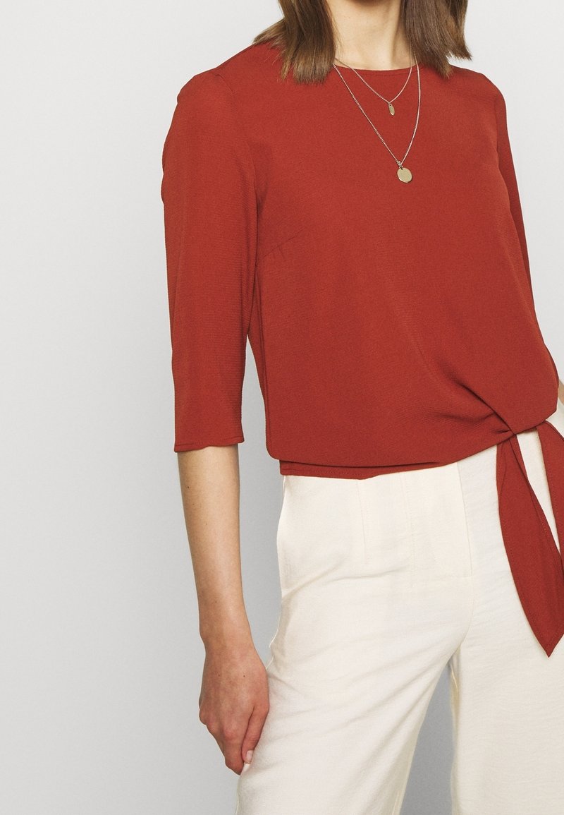 Femme portant un haut couleur rouille à manches trois-quarts avec un nœud sur le côté et un pantalon blanc taille haute, portant deux colliers pendentifs en or.