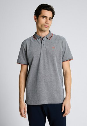 Mann steht und trägt ein graues Poloshirt mit kurzen Ärmeln, rotem und schwarzem Besatz am Kragen und an den Ärmeln, kombiniert mit dunkler Hose.