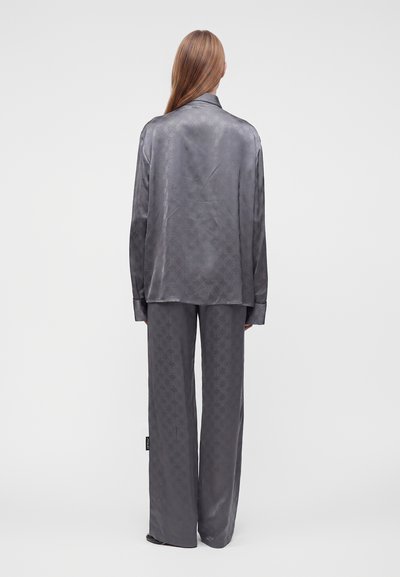 Personne aux longs cheveux portant un ensemble de pyjama soyeux gris ample, vue de dos sur un fond clair uni.