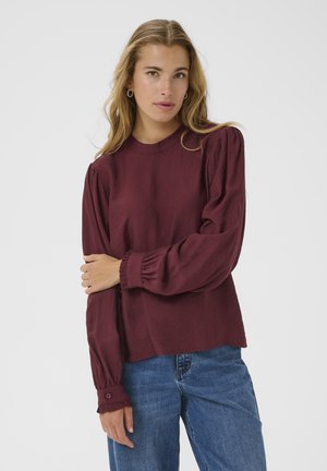 Blusa bordeaux a maniche lunghe con spalle a palloncino e polsini arricciati, realizzata in tessuto leggero. Abbinata a jeans blu a vita alta.
