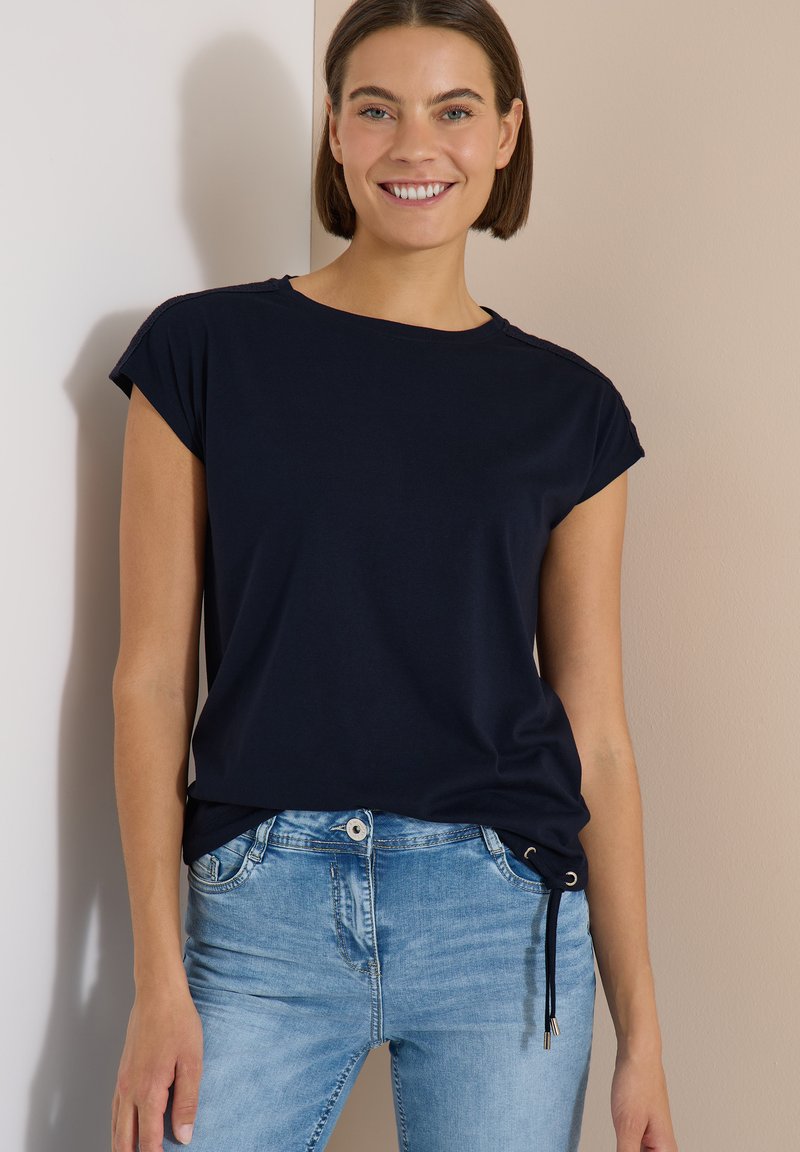 Femme souriante aux cheveux courts bruns portant un haut noir à manches courtes et un jean bleu clair, debout contre un mur beige.