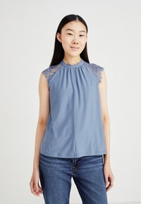 Top blu senza maniche con scollo alto, caratterizzato da inserti in pizzo trasparente sulle spalle e una parte anteriore plissettata. Abbinato a jeans in denim.