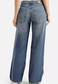 Osoba w niebieskich jeansach z wysokim stanem i szerokimi nogawkami, z dwiema tylnymi kieszeniami, stojąca na tle białym.