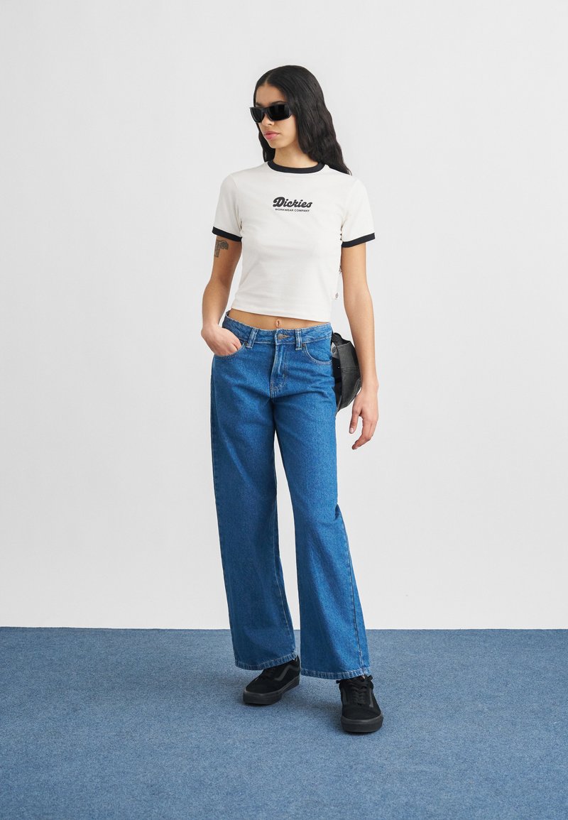 Femme portant des lunettes de soleil noires, un t-shirt court blanc Dickies, un jean large bleu, des baskets noires, debout sur un sol recouvert de moquette bleue.