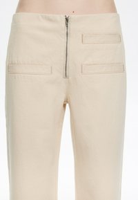 Pantalones de algodón color beige con cintura ajustada, cierre de cremallera en la parte delantera y dos bolsillos traseros con acentos de costura horizontal.