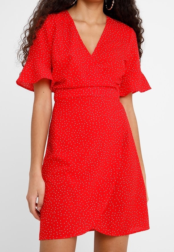 Femme portant une robe portefeuille rouge à pois blancs, avec de courtes manches évasées et un décolleté en V, debout devant un fond uni.