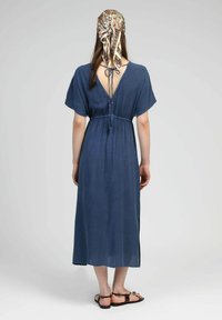 Robe maxi fluide en tissu bleu marine avec des manches courtes. Elle présente un dos ouvert noué, une taille élastique et des fentes latérales pour plus de mouvement.