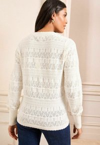 White knitted sweater featuring intricate openwork patterns, a textured ribbed yoke, and scalloped cuffs, paired with dark blue jeans.