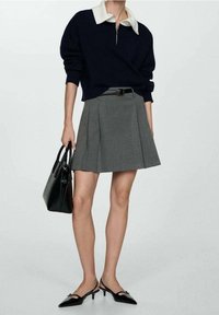 Suéter de punto azul marino con un cuello blanco, falda de tablas gris, bolso negro y zapatos de tacón negros con una correa sobre el pie.