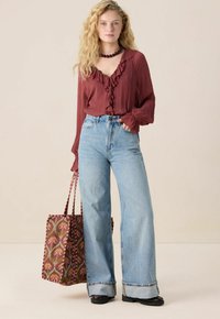 Blouse bourgogne avec encolure à volants, jeans bleu clair à jambes larges et sac fourre-tout à motifs maron et verts.
