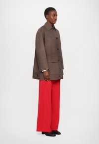 Jachetă oversized maro cu guler, închidere cu butoane și buzunare frontale, purtată cu pantaloni largi roșii aprinși și pantofi negri.